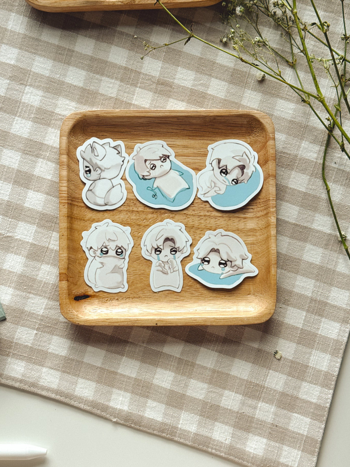 Six love and deepspace love interest chibi stickers displayed on a small wooden tray over a beige-and-white checkered cloth, with dried flowers and part of another tray visible around it.